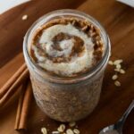 This is a photo of cinnamon roll overnight oats with a spoon and cinnamon sticks.