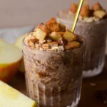 a glass cup with apple pie overnight oats with a spoon in it and an apple on the side.