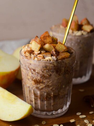 a glass cup with apple pie overnight oats with a spoon in it and an apple on the side.