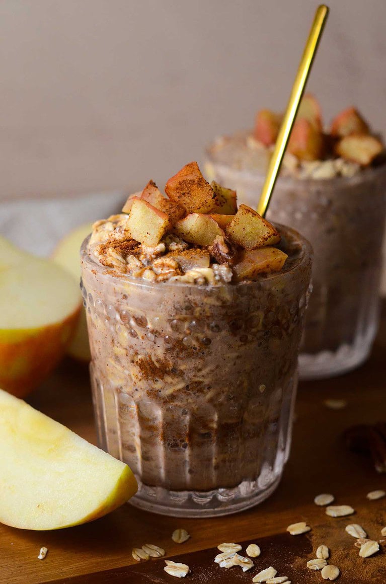 a glass cup with apple pie overnight oats with a spoon in it and an apple on the side.