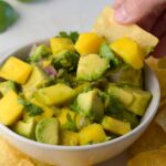 mango avocado salsa in a bowl with a hand dipping a chip in it.