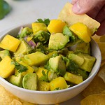 mango avocado salsa in a bowl with a hand dipping a chip in it.