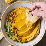 pumpkin hummus in a bowl with a hand dipping pita into it.