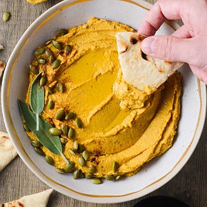 pumpkin hummus in a bowl with a hand dipping pita into it.