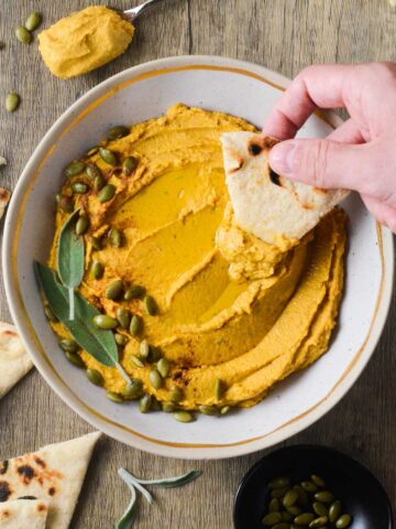 pumpkin hummus in a bowl with a hand dipping pita into it.