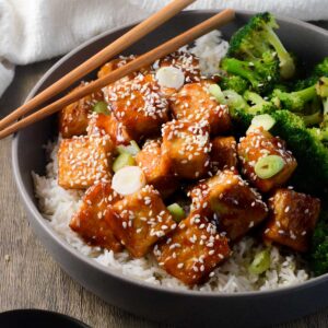 bowl of sticky tofu with chopsticks, rice, and broccoli.