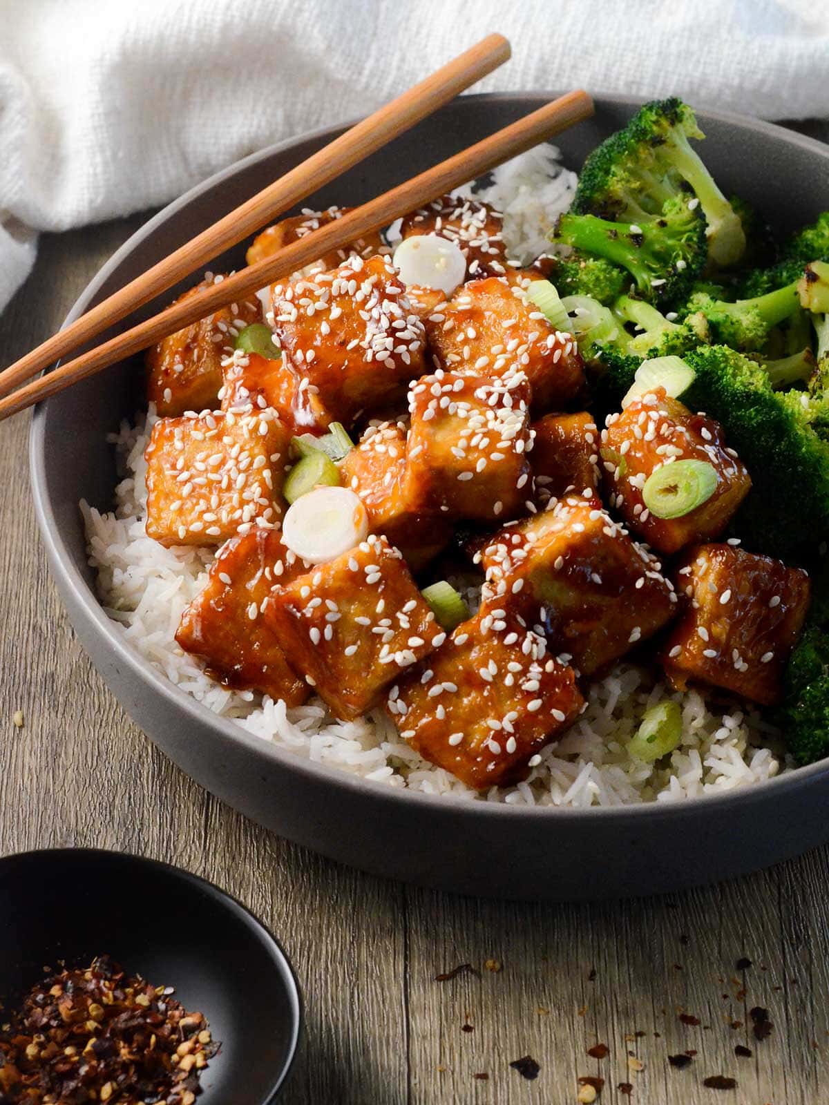 bowl of sticky tofu with chopsticks, rice, and broccoli.