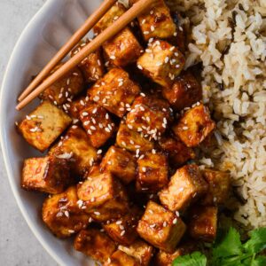 crispy asian tofu in a bowl with brown rice, cilantro, and chopsticks.