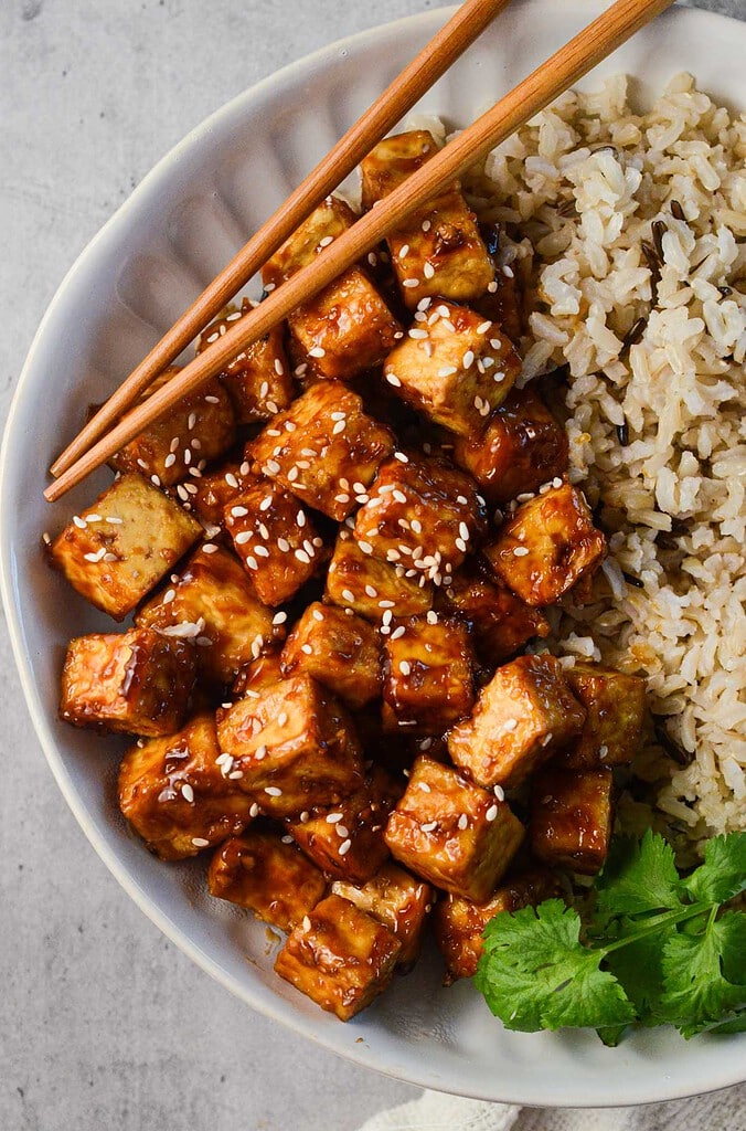 crispy asian tofu in a bowl with brown rice, cilantro, and chopsticks.