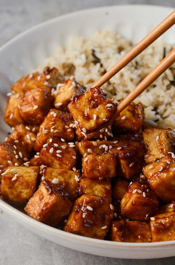 crispy asian tofu with sesame seeds on top and a chopstick picking it up.
