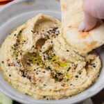 everything bagel hummus in a shallow bowl with a hand dipping pita bread in it.
