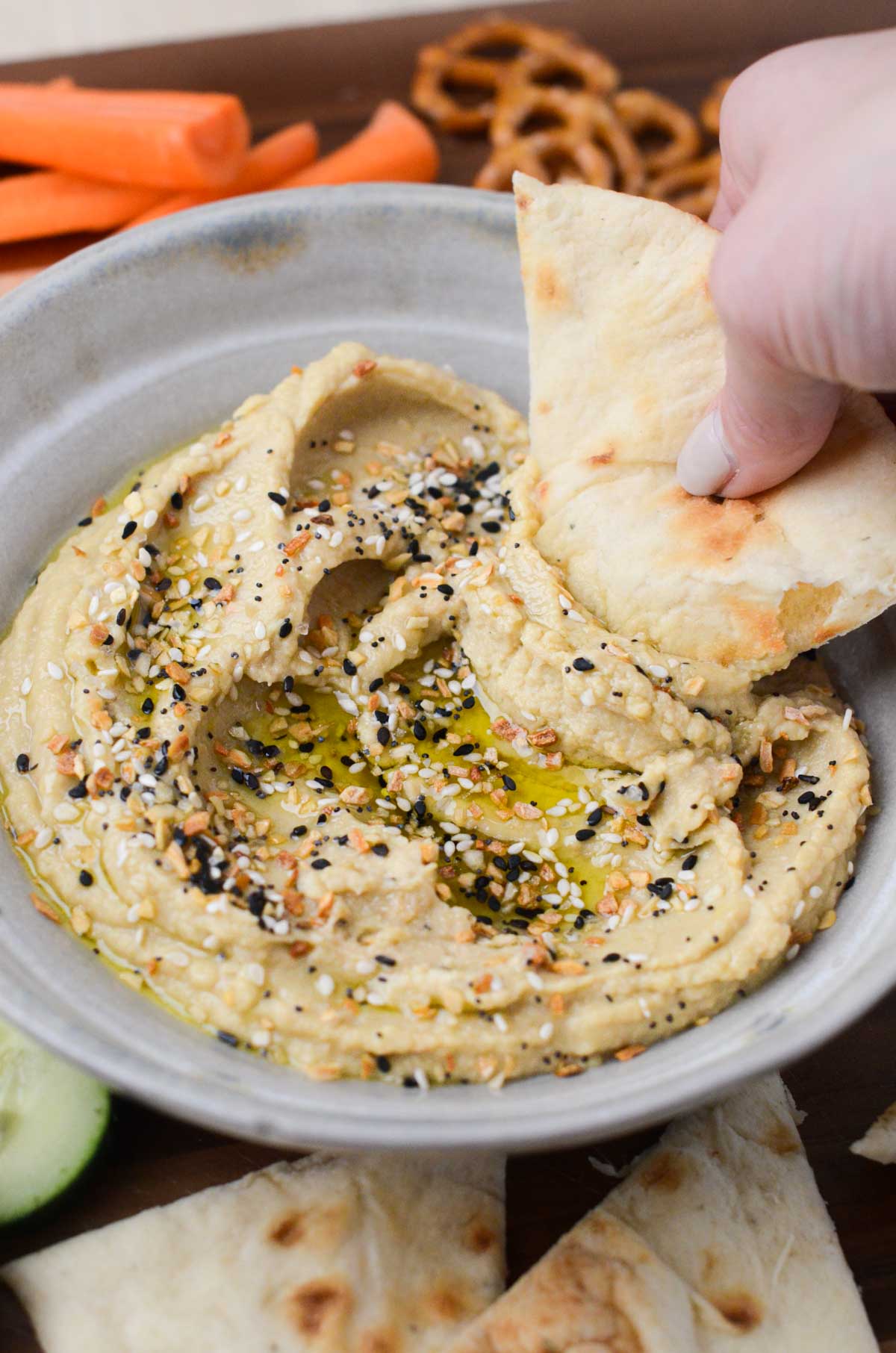 everything bagel hummus in a shallow bowl with a hand dipping pita bread in it.