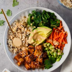 asian tofu buddha bowl surrounded by carrots, brown rice, sauce, and cilantro.