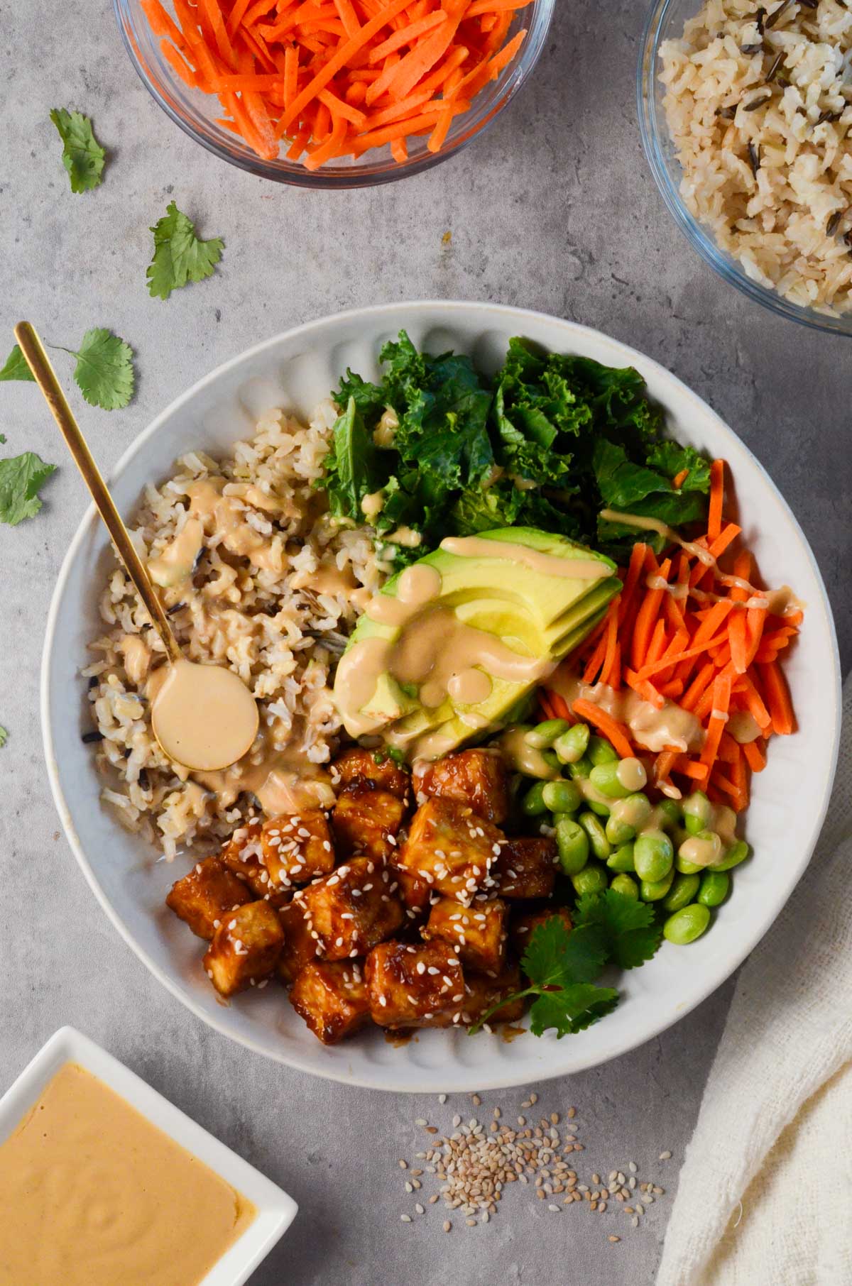 asian tofu buddha bowl surrounded by carrots, brown rice, sauce, and cilantro.